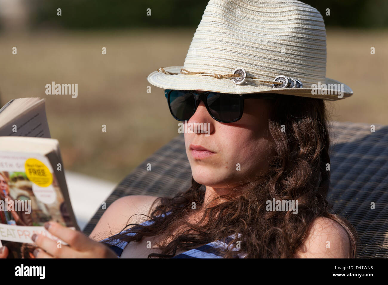 Young woman in bikini, lunettes de soleil et chapeau, se détend en dehors au soleil sur une chaise longue inclinable comme elle lit un livre de poche. Banque D'Images