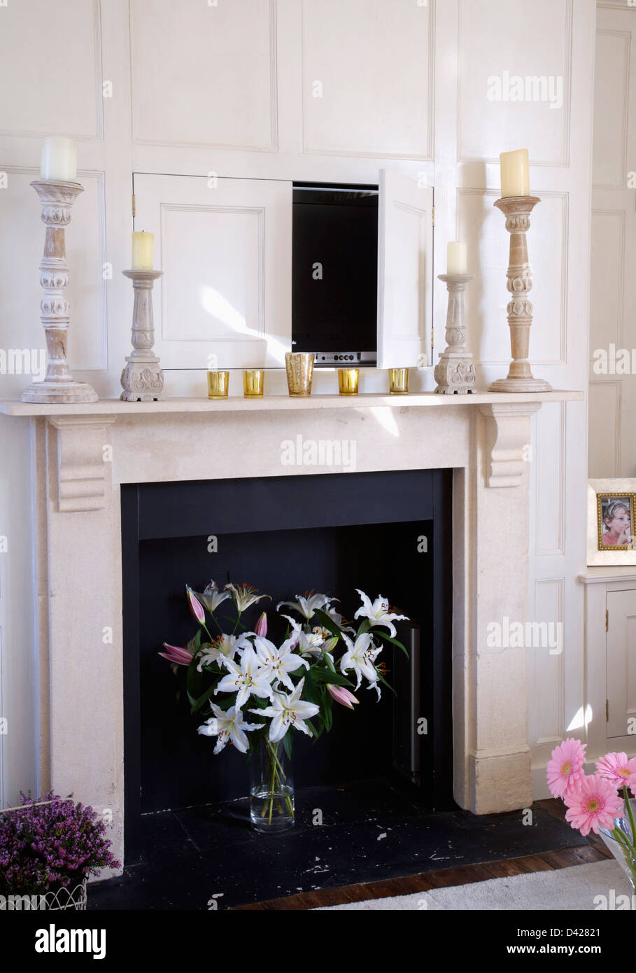 Porte ouverte à la télévision dissimulé dans des bois au-dessus de cheminée en marbre avec vase de lys blancs dans la grille Banque D'Images