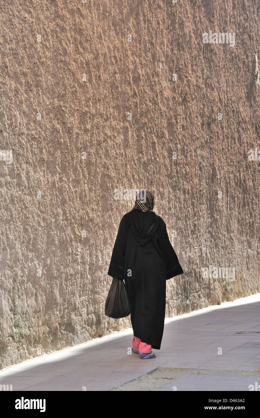 Vue arrière de la femme marocaine en manteau noir et voile le long mur de briques élevé dans la ville de la côte atlantique d'Essaouira. Banque D'Images