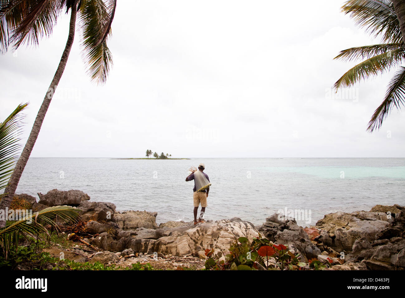 Un pêcheur tenant un filet se dresse en bordure de rochers surplombant l'océan sur un jour nuageux. Banque D'Images