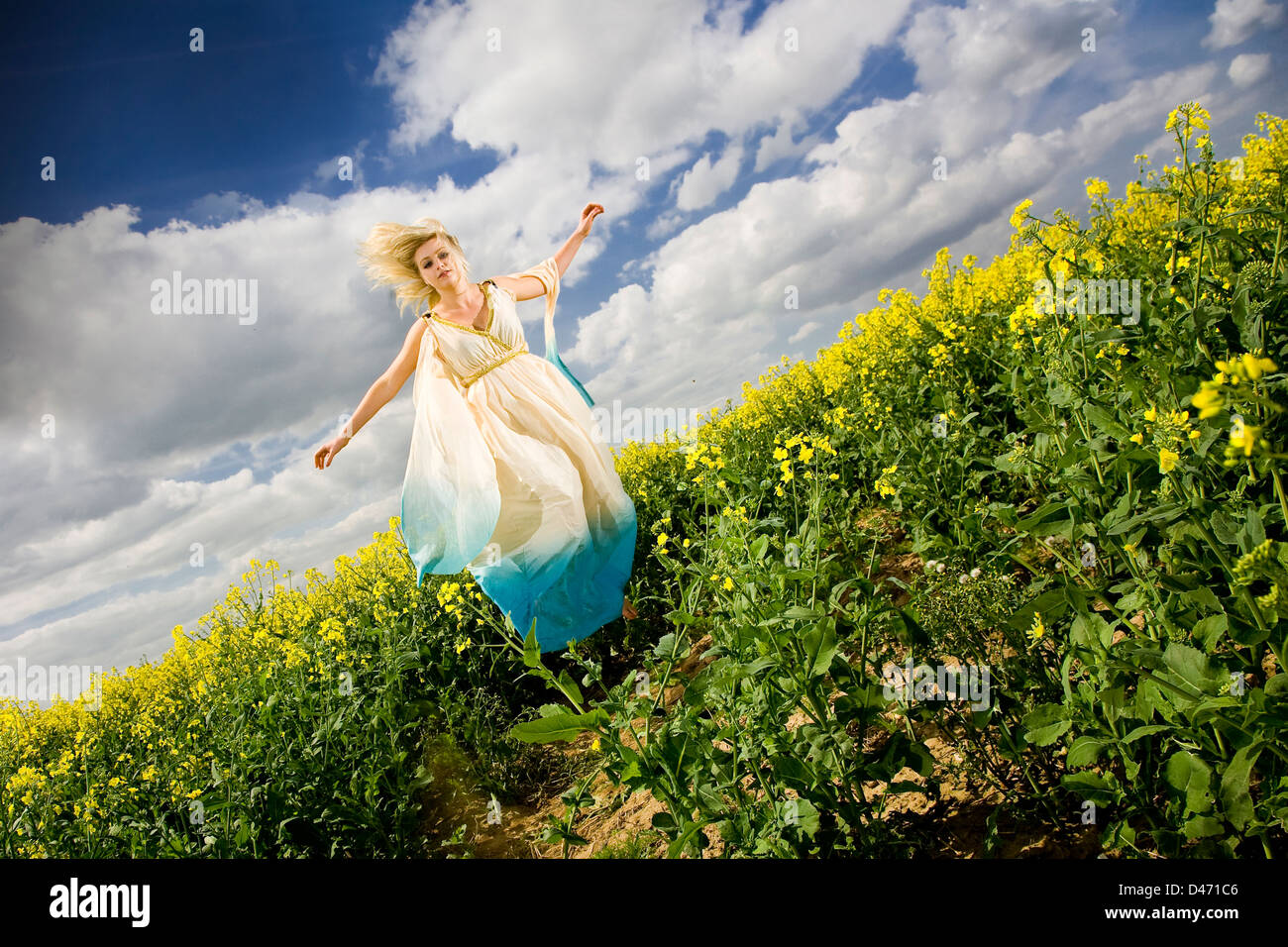 Jeune femme blonde en robe de style grec sarcelle crème sautant au-dessus de champs de colza jaune avec ciel bleu et nuages Banque D'Images