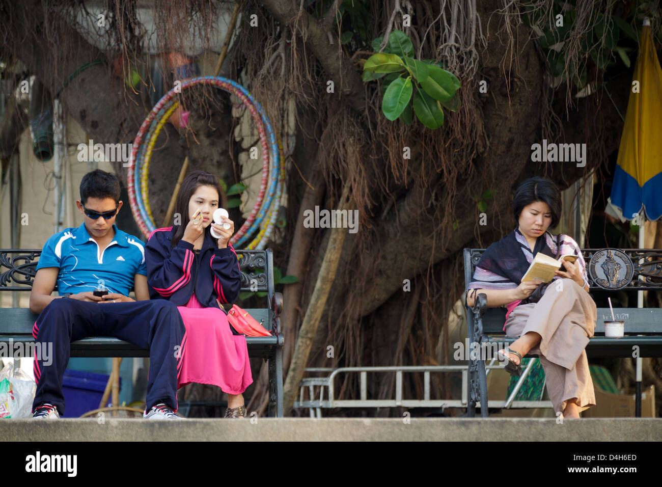 Les thaïs assis dans un parc, l'un est l'utilisation de smartphone, l'un est l'application de rattraper , le dernier est lecture bookd, Bangkok, Thaïlande Banque D'Images