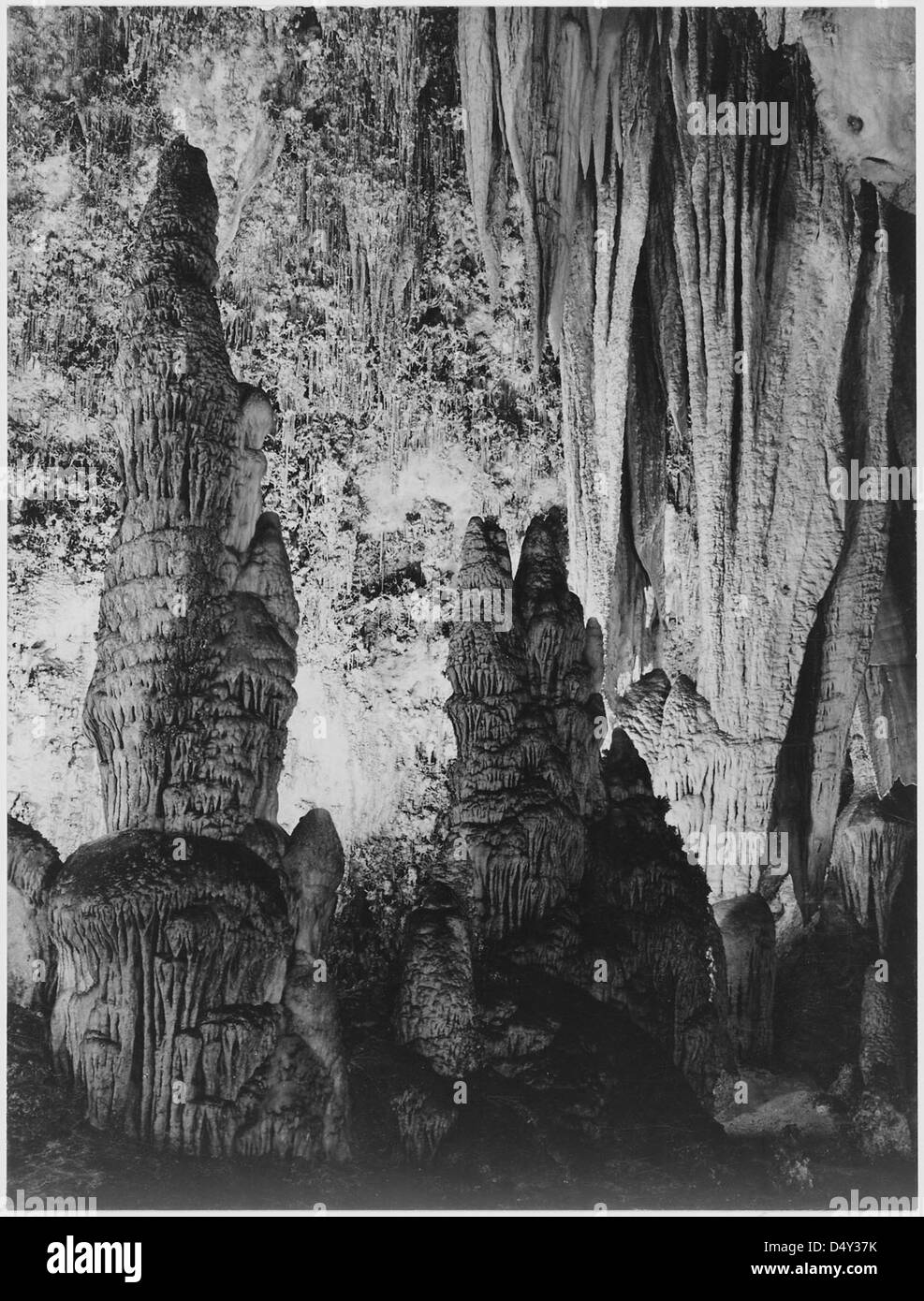 La photographie d'Ansel Adams capture les spectaculaires formations de stalagmites et les rideaux d'onyx à l'intérieur du Kings Palace dans le parc national de Carlsbad Caverns, au Nouveau-Mexique. L'image montre la grandeur de ces structures minérales naturelles en noir et blanc, soulignant les détails complexes et les textures des formations de la grotte. Banque D'Images