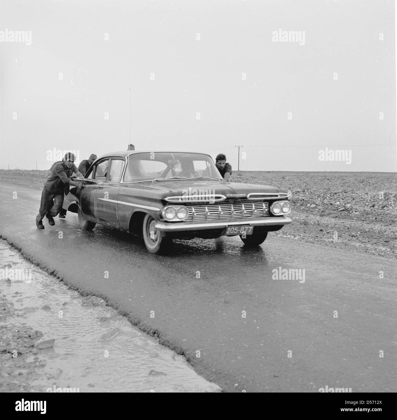 Israël, années 1950. Tableau historique des quatre hommes poussant une vieille voiture américaine le long d'une piste ou sur route goudronnée dans un désert aride comme domaine. Banque D'Images