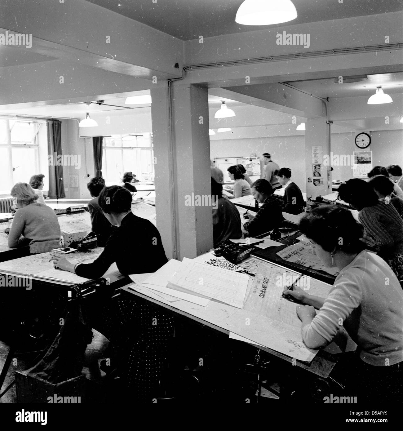 Image historique, années 1960. Groupe de femmes travaillant dans un bureau de type industriel sur de grands bureaux en métal pour établir les chiffres de vente. Banque D'Images