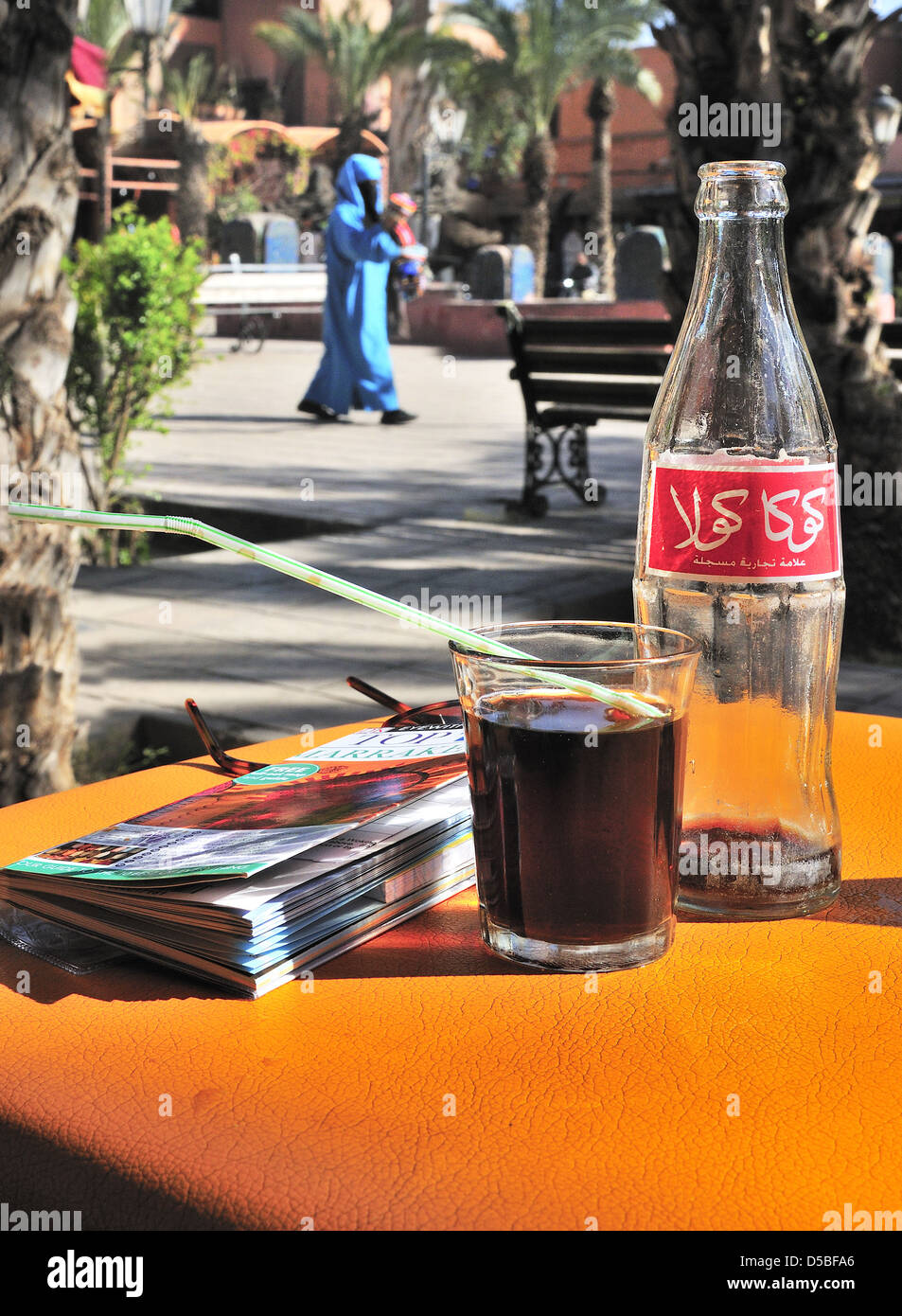 Table dans un café avec le guide de Marrakech, bouteille de Coca Cola arabe et verre plein et dame marocaine traversant la piazza à Marrakech, Maroc Banque D'Images