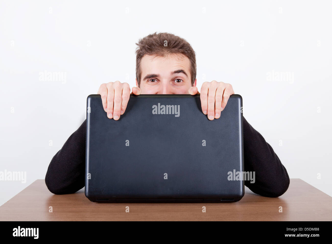 Jeune homme de se cacher derrière un ordinateur portable sur un bureau Banque D'Images