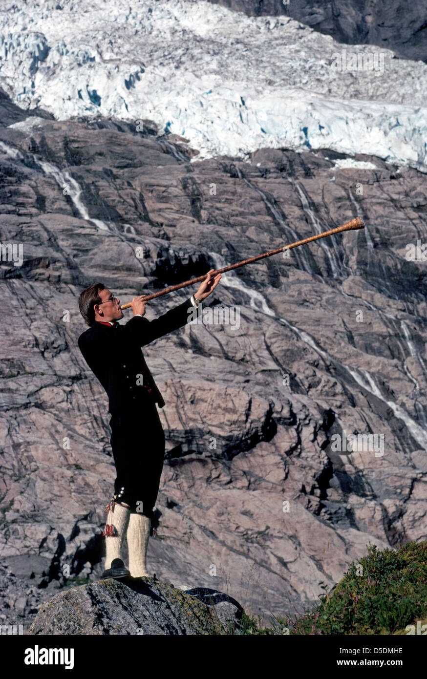 Un jeune homme norvégien en vêtements traditionnels d'antan souffle un cor des alpes en bois vintage sous un glacier de montagne à Fjaerland, la Norvège. Banque D'Images