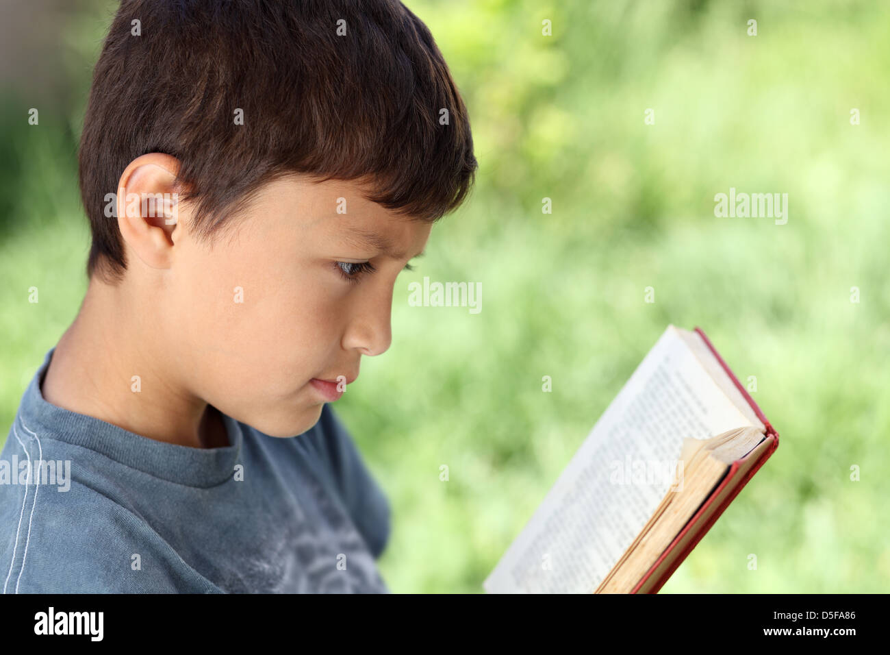 Young boy reading book extérieur avec fond vert naturel et profondeur de champ - avec copie espace à droite Banque D'Images