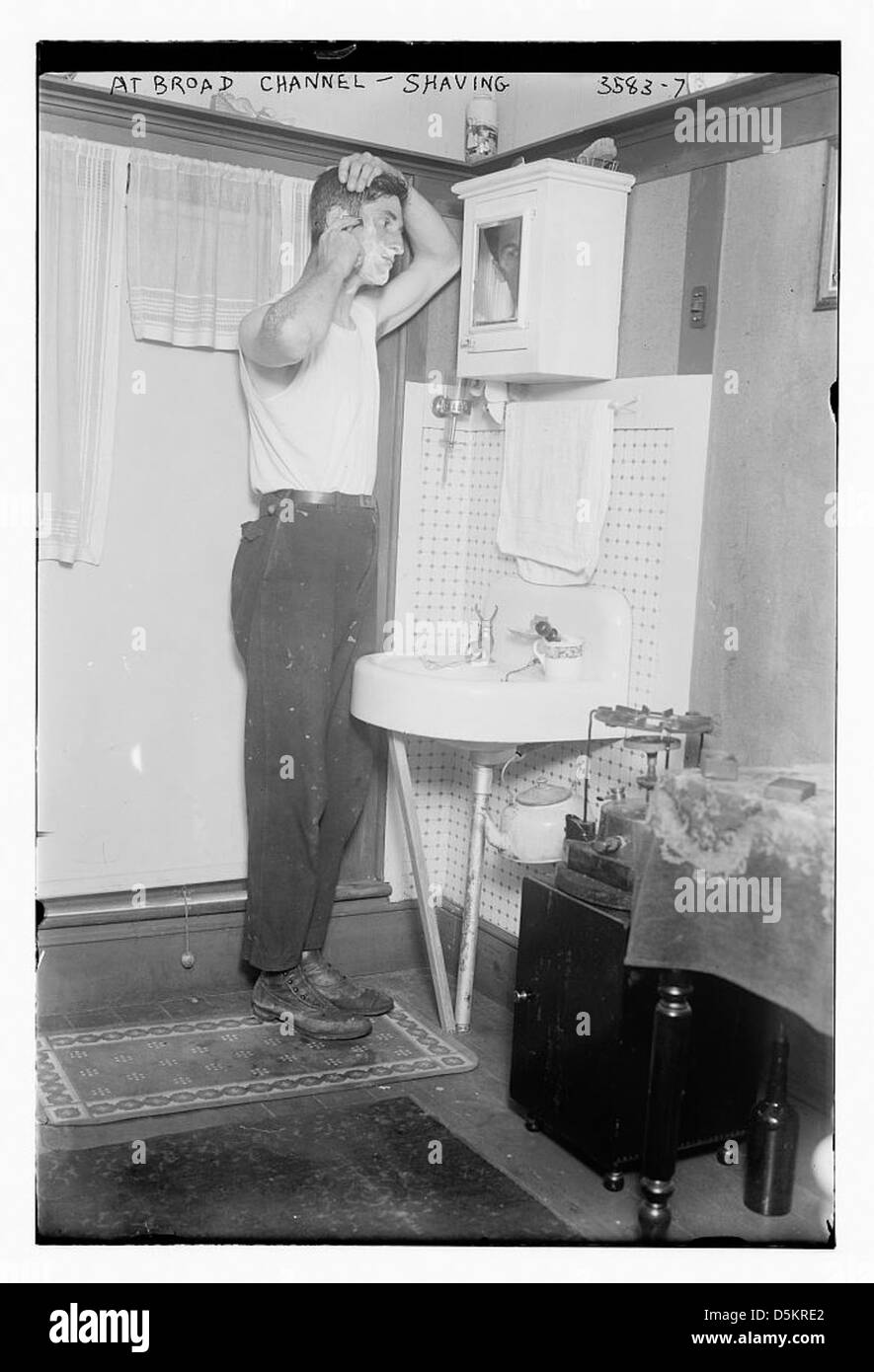 Cette photographie montre un homme se rasant dans une salle de bain à Broad Channel, Queens, New York. La scène capture la vie quotidienne dans le quartier avec l'homme debout devant un évier, à l'aide d'un miroir. Banque D'Images