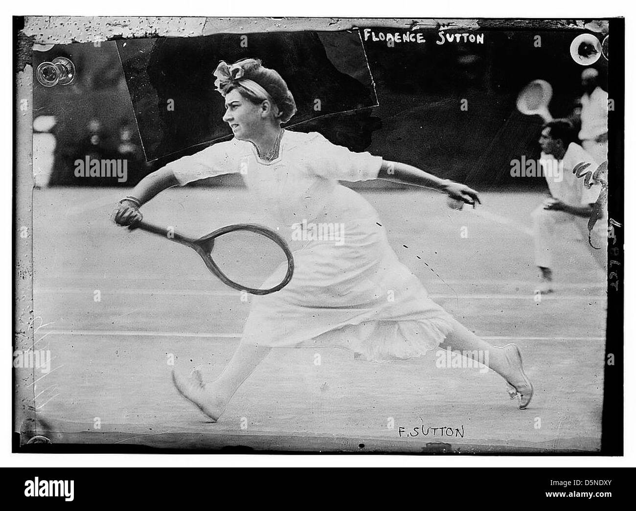 Florence Sutton est capturée dans un tir d'action de tennis des années 1910, montrant un coup de revers tout en portant une tenue appropriée d'époque. L'image en noir et blanc met en valeur sa forme sportive et sa grâce sportive. Banque D'Images