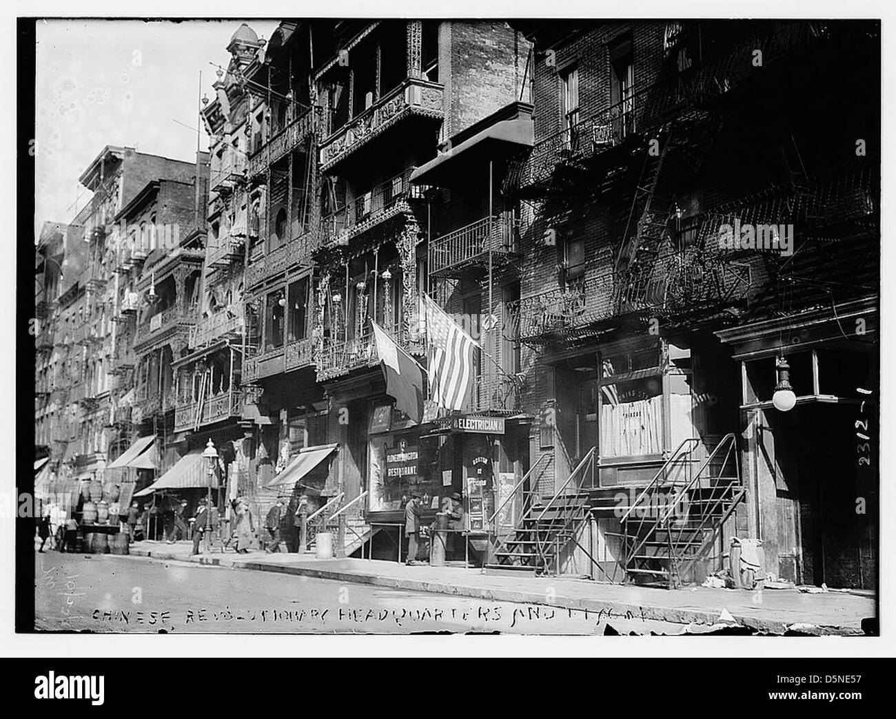 Cette image représente une scène de rue sur Mott Street, New York, possiblement liée à un quartier général révolutionnaire chinois. La scène présente des bâtiments, des entreprises telles que Flower Kingdom Restaurant, et un quartier avec des appartements et des tonneaux, reflétant la vie du Chinatown au début du XXe siècle et les activités révolutionnaires en Amérique. Banque D'Images