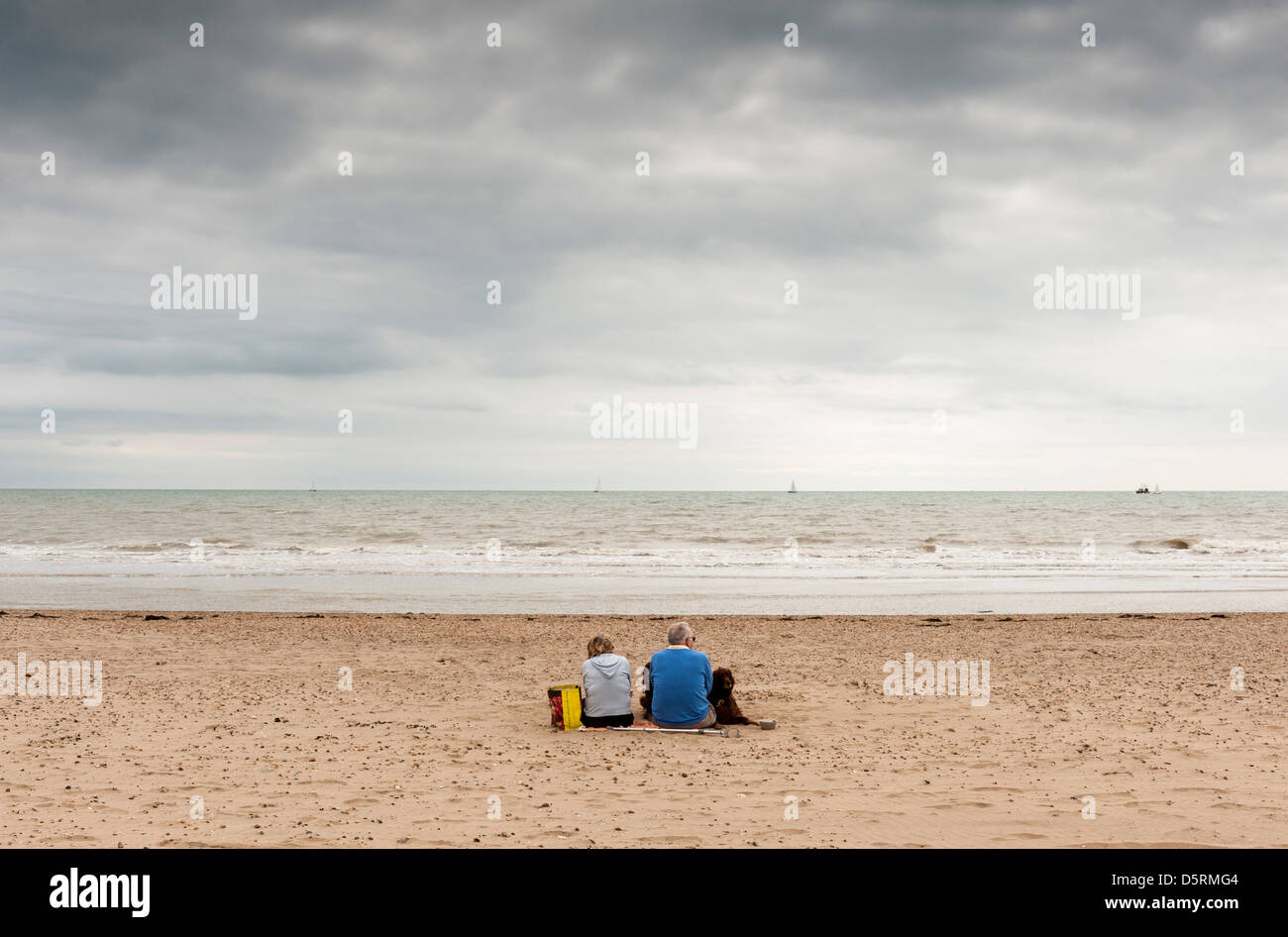 Un couple et leur chien assis sur la plage à Camber Sands, East Sussex, England, UK - comme les nuages de pluie gris recueillir Banque D'Images