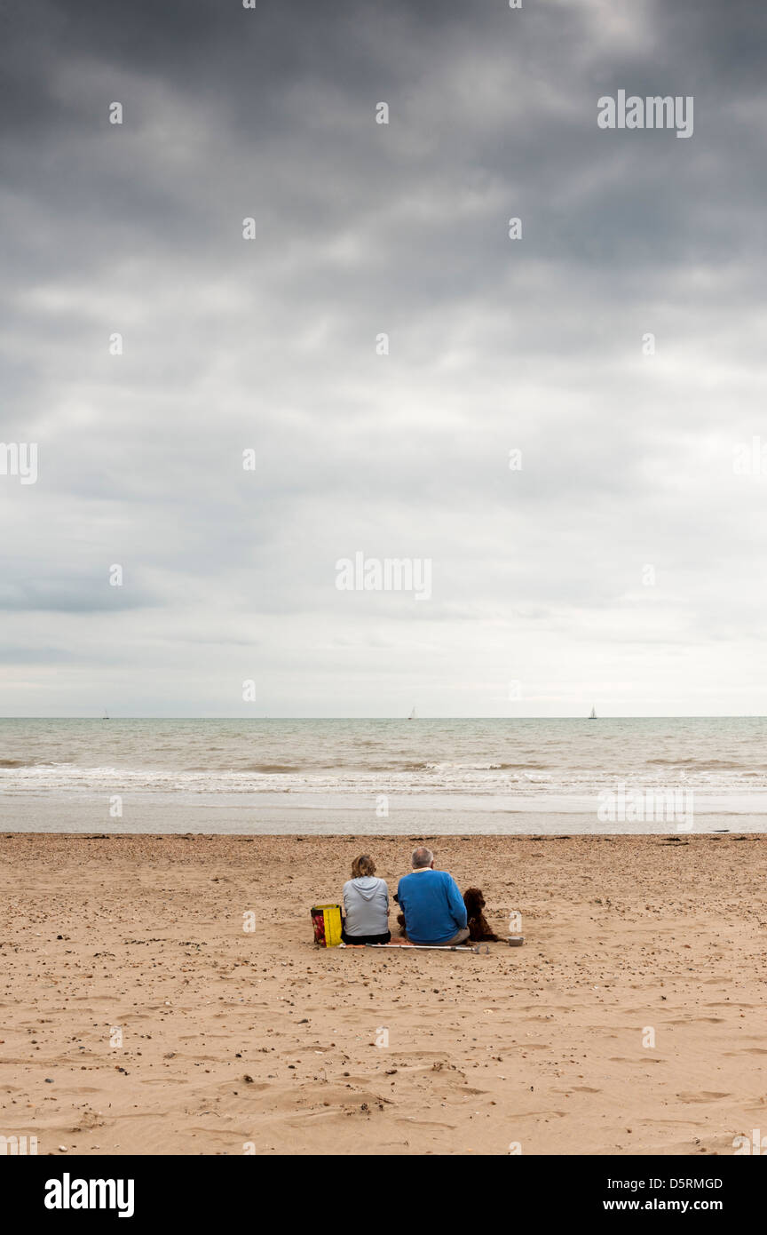 Couple et leur chien assis sur la plage de Camber Sands, East Sussex, alors que les nuages de pluie s'amoncellent Banque D'Images