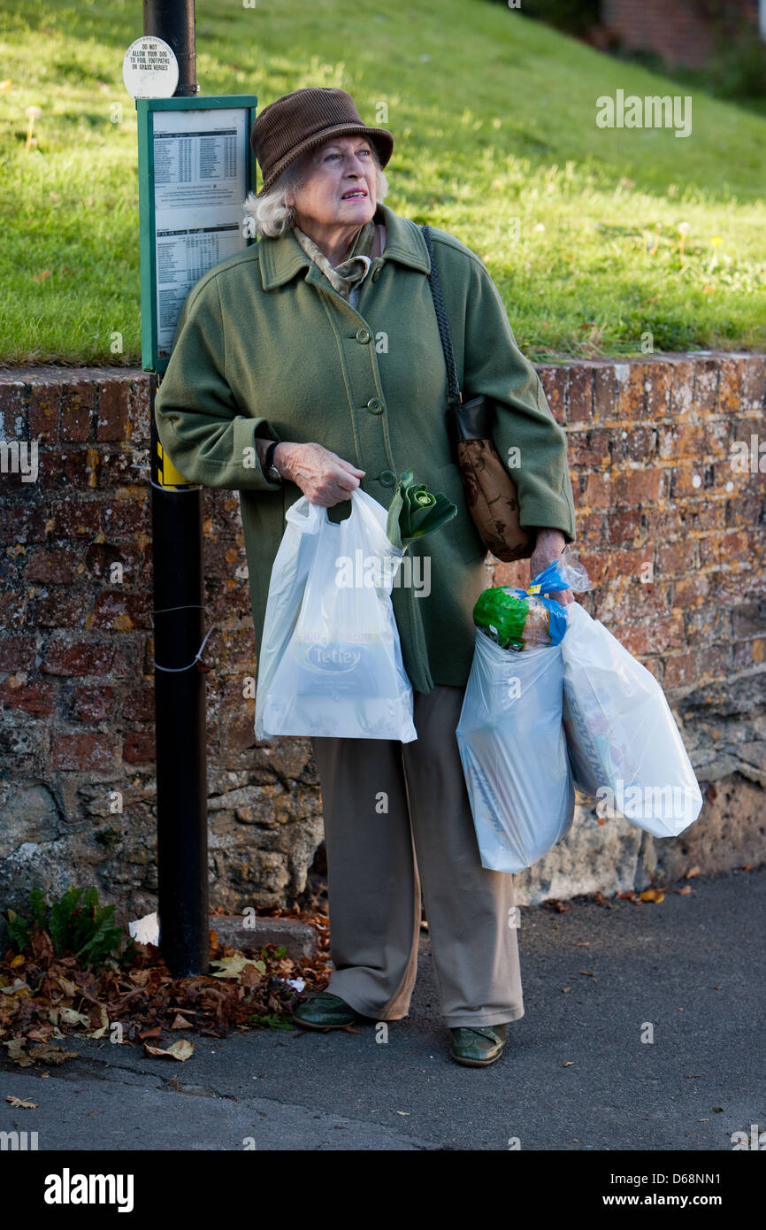 Femme âgée en attente à un arrêt de bus avec des sacs de shopping. Banque D'Images