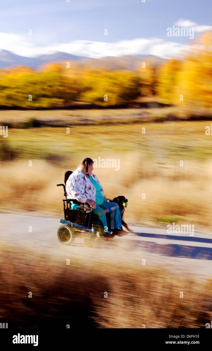 Femme handicapée d'âge moyen conduire son fauteuil roulant motorisé vers le bas une petite ville sentier de marche Banque D'Images