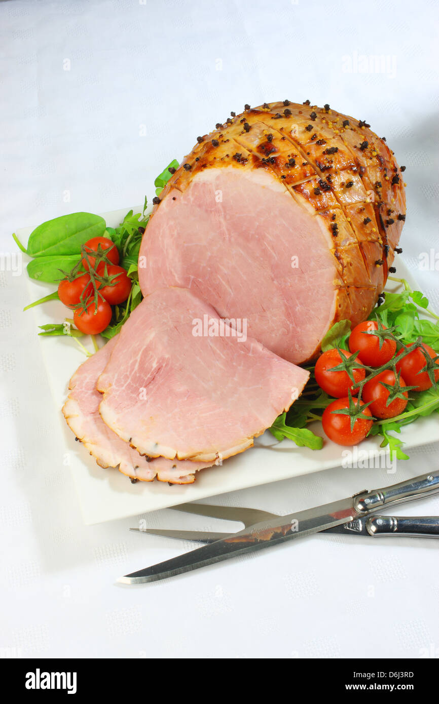 Jambon cuit au four avec des clous de girofle et les tomates cerises et couteau et fourchette sur un carré blanc sur la plaque d'une nappe blanche Banque D'Images