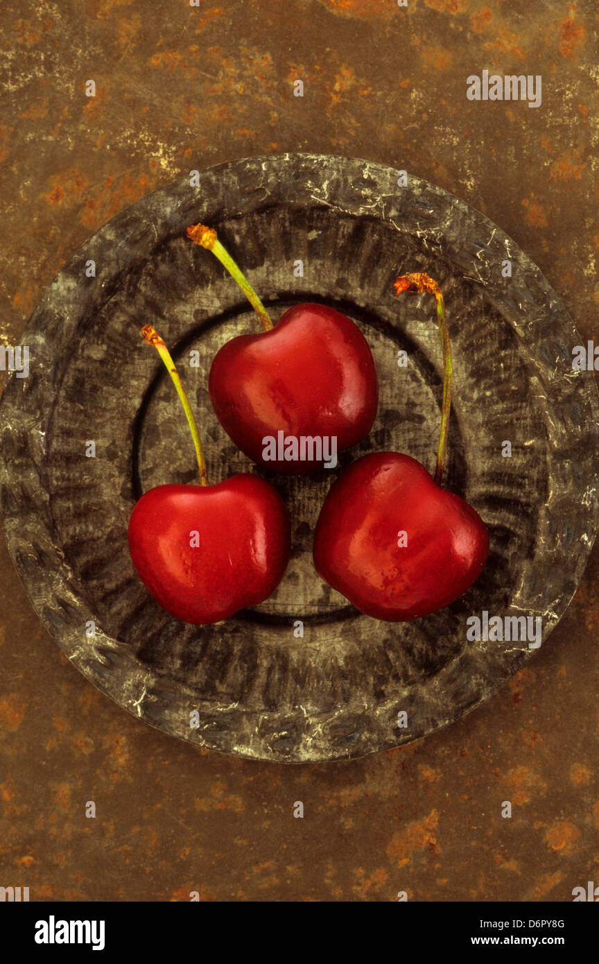 Trois cerises rouge foncé avec des tiges vertes situées dans de petits plats d'étain ornée sur feuille de métal rouillé Banque D'Images