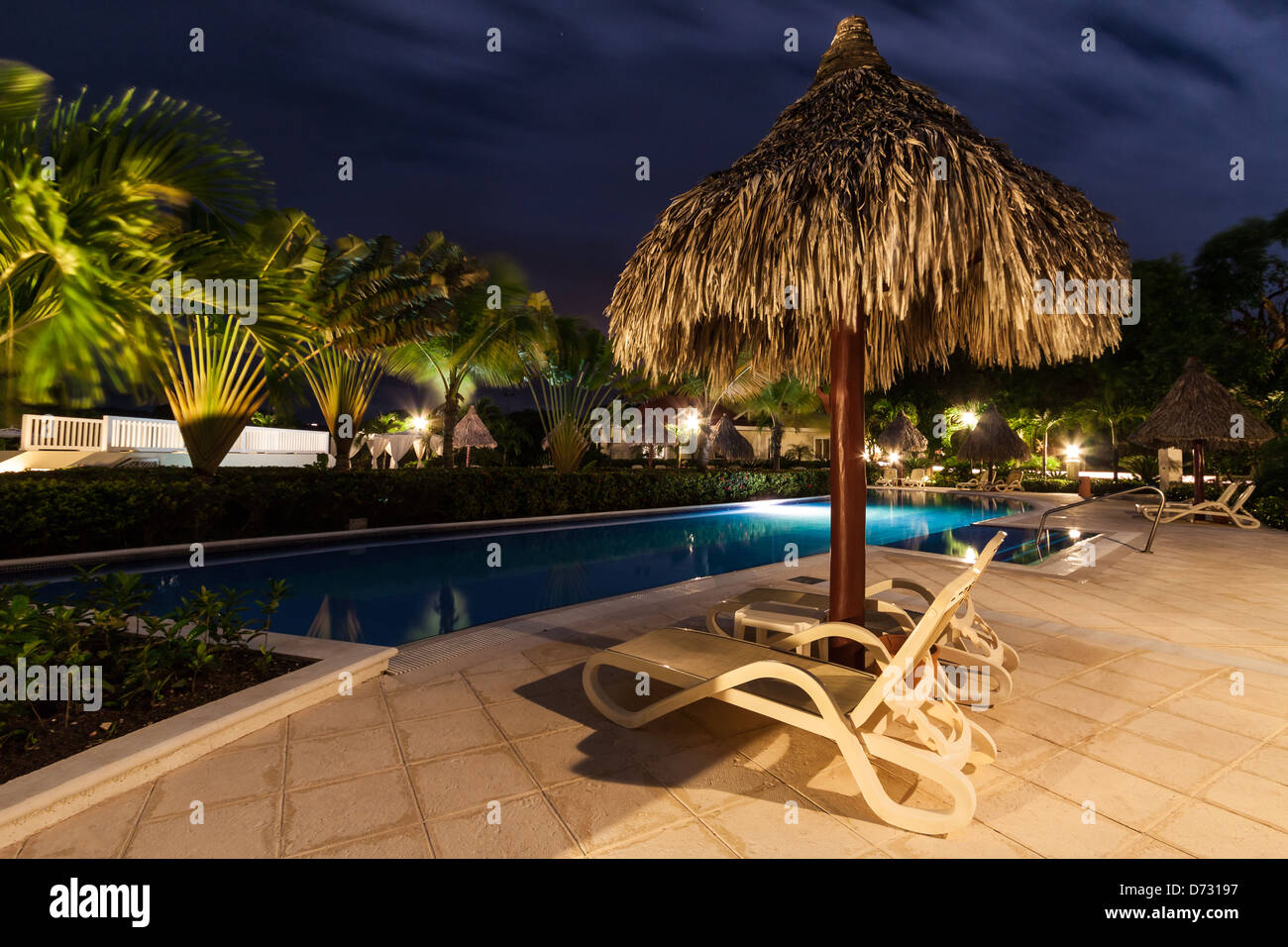 Parapluie de paille et deux sièges de plage à proximité d'une piscine illuminée par nuit Banque D'Images