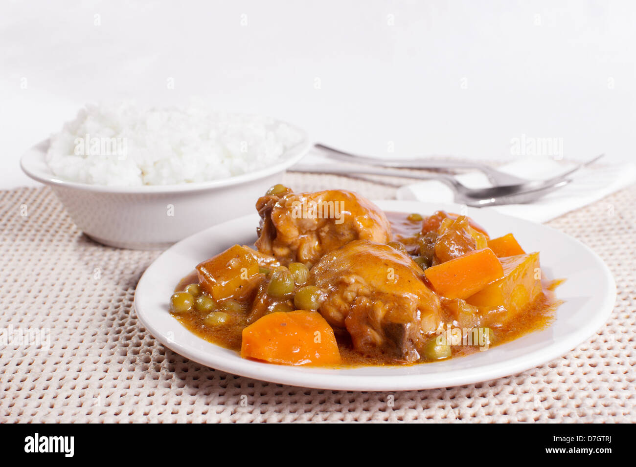 Ragoût de poulet avec pommes de terre, carottes, petits pois à la sauce tomate. bol de riz vapeur, fourchette et cuillère en arrière-plan. Banque D'Images