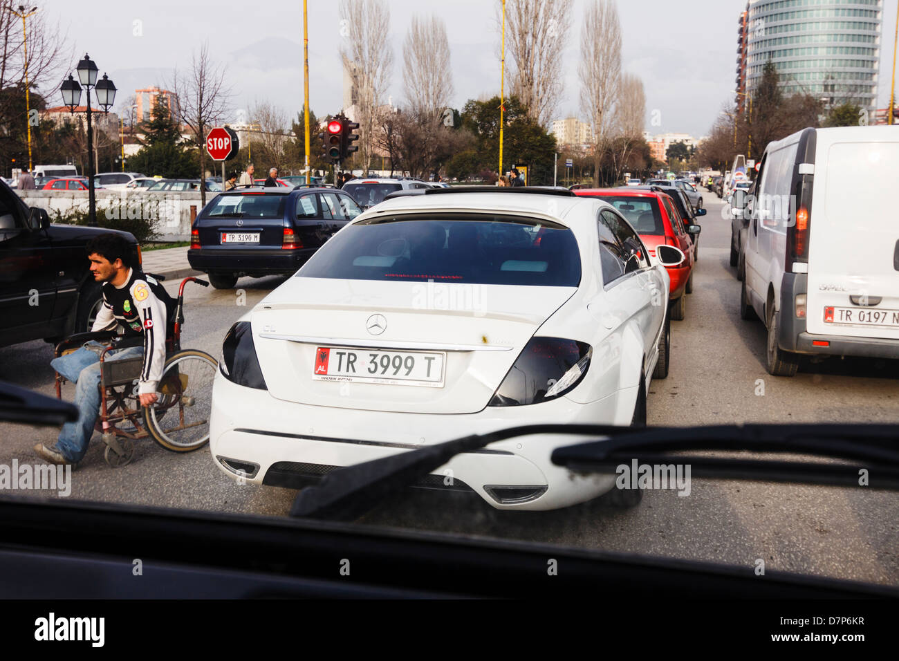 Homme handicapé sur fauteuil roulant en passant par un embouteillage avec des voitures de luxe dans le centre de Tirana, Albanie Banque D'Images