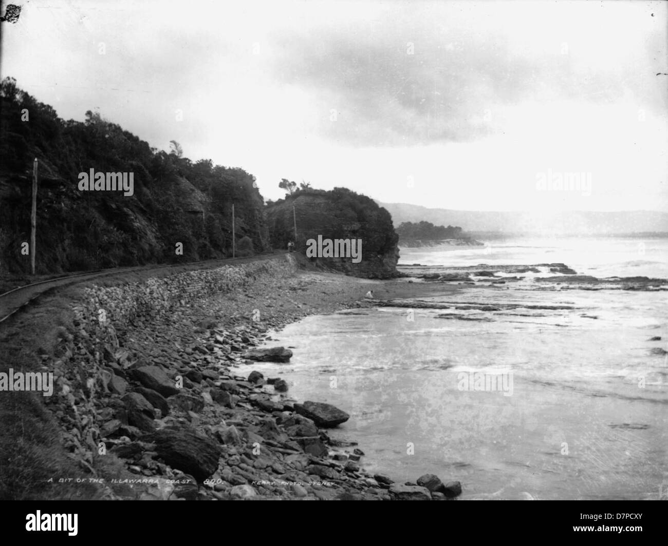 Cette photographie historique de Charles H. Kerry capture la côte d'Illawarra, mettant en valeur North Wollongong Beach et le paysage côtier environnant. L'image comprend le port de Wollongong, avec des voies de chemin de fer ou de tramway visibles, et une représentation globale de l'infrastructure et de la beauté naturelle de la région au début du XXe siècle. Banque D'Images
