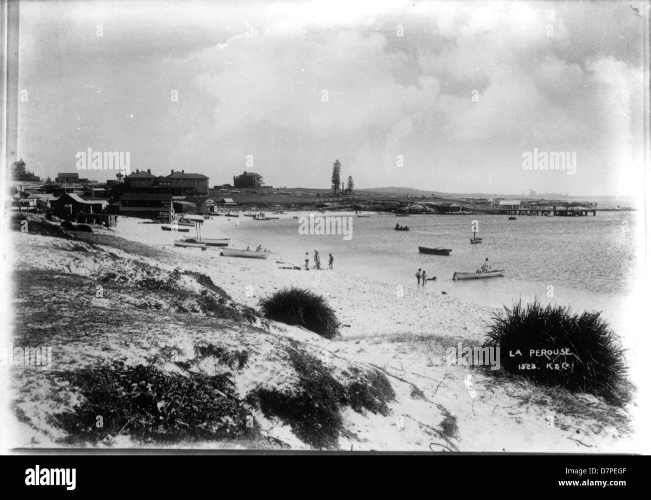 Une photographie en noir et blanc de la région de la Pérouse dans la baie de Frenchmans, avec une plage, des bateaux, des bâtiments et des gens au bord du rivage. L'image met en évidence le paysage tranquille, avec des nageurs et la vie végétale visibles le long du rivage, capturant un moment dans l'histoire de la région. Banque D'Images