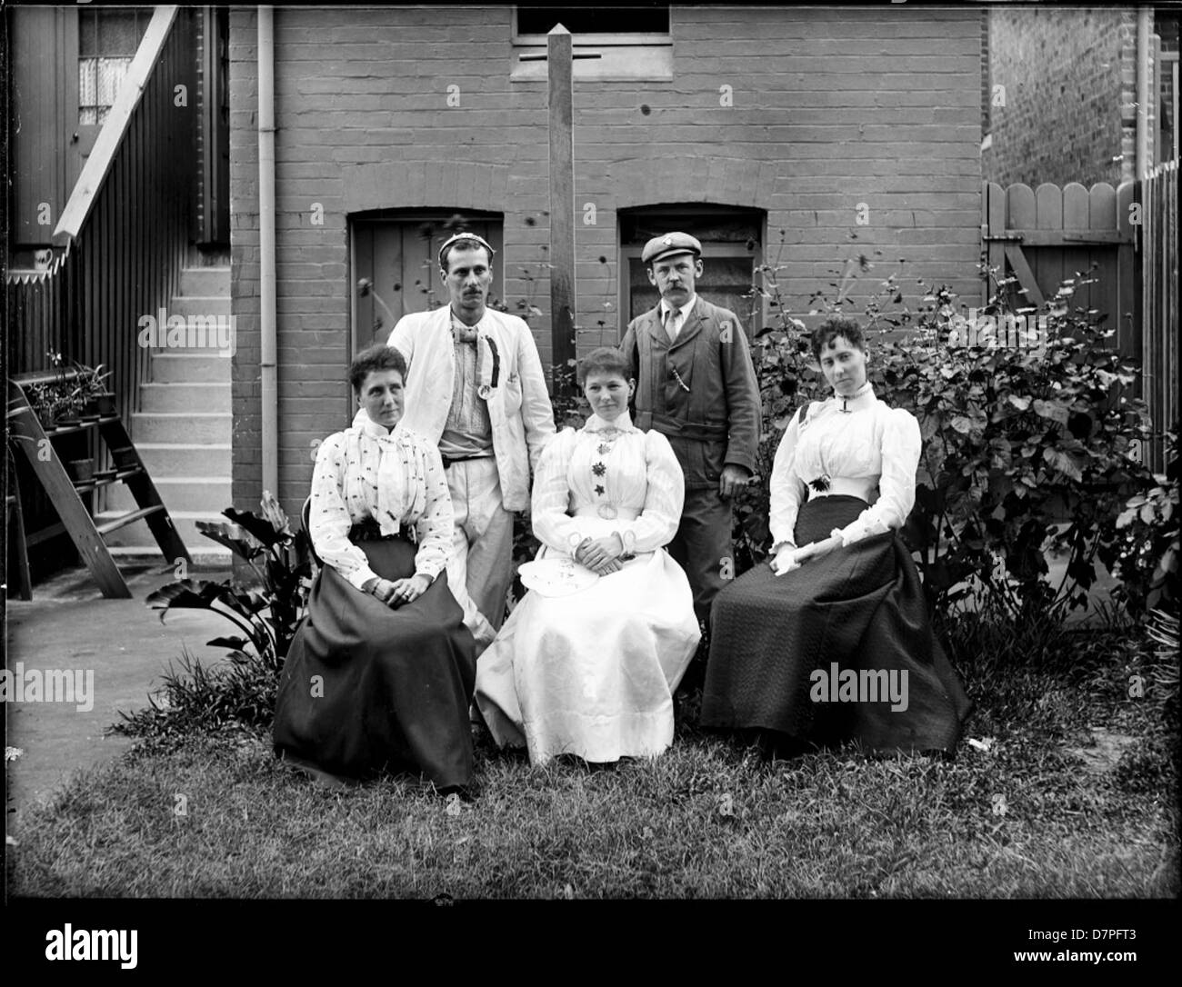 Une photographie de 1890-1900, capturant trois femmes et deux hommes dans le jardin d'une maison en terrasse. L'image reflète la mode et la vie sociale de la fin du XIXe siècle, mettant en valeur une tenue habillée, un jardin bien entretenu et un aperçu du mode de vie de cette période. Le cadre comprend une clôture décorative, une porte formelle et des éléments fantaisistes comme des arbustes et des escaliers. Banque D'Images