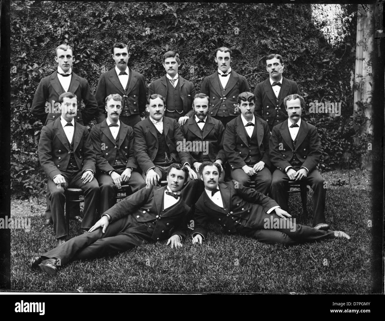 Cette photographie historique montre un groupe de 13 jeunes hommes en costumes sombres, posant pour l'appareil photo. L'image, qui fait partie de la collection du Powerhouse Museum, offre un aperçu de la mode et des dynamiques sociales du début du XXe siècle. Banque D'Images