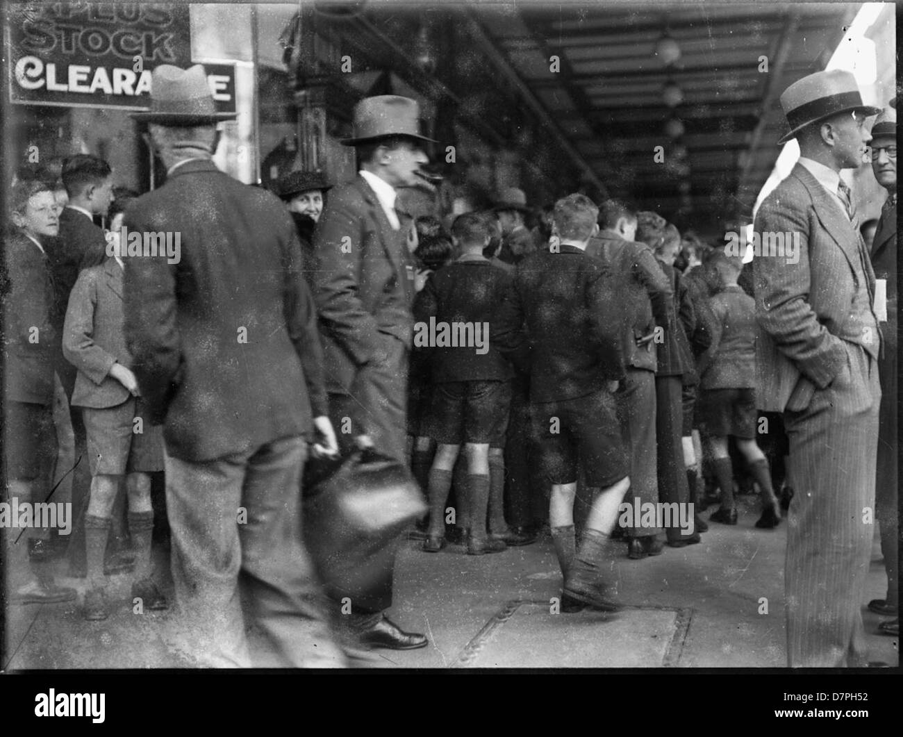 Cette image représente un groupe d'écoliers et un homme à l'extérieur du grand magasin F.J. Palmer & sons, capturés dans un moment historique. La scène reflète la vie quotidienne au début du XXe siècle, avec des gens rassemblés devant le magasin, peut-être en attente de transport. Le cadre se trouve à proximité d'une gare, révélateur du commerce et du système de transport animés de l'époque. Banque D'Images