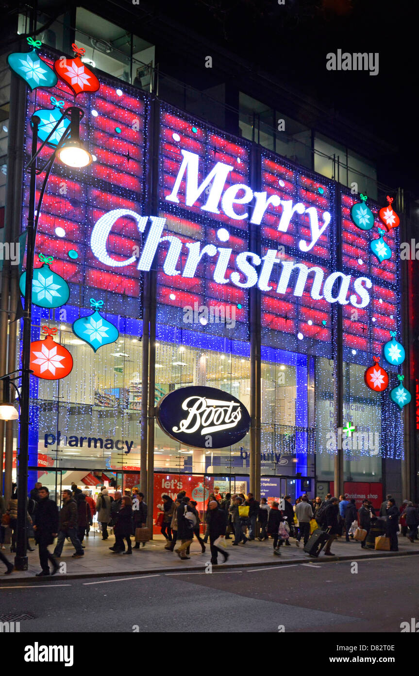 Les amateurs de shopping de nuit dans la rue devant les vitrines du magasin  Boots Chemist & Magasin de pharmacie Oxford Street grand signe Joyeux Noël  dans les lumières Londres Angleterre Royaume-Uni