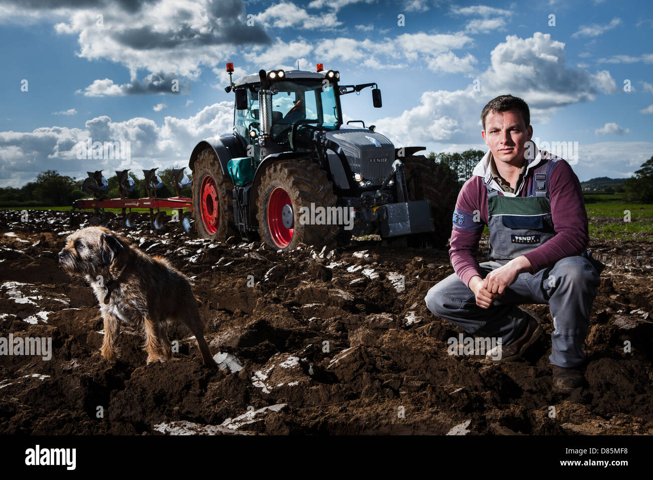 Portrait d'un jeune agriculteur et son petit chien en avant d'un tracteur et charrue dans le champ labouré. Banque D'Images