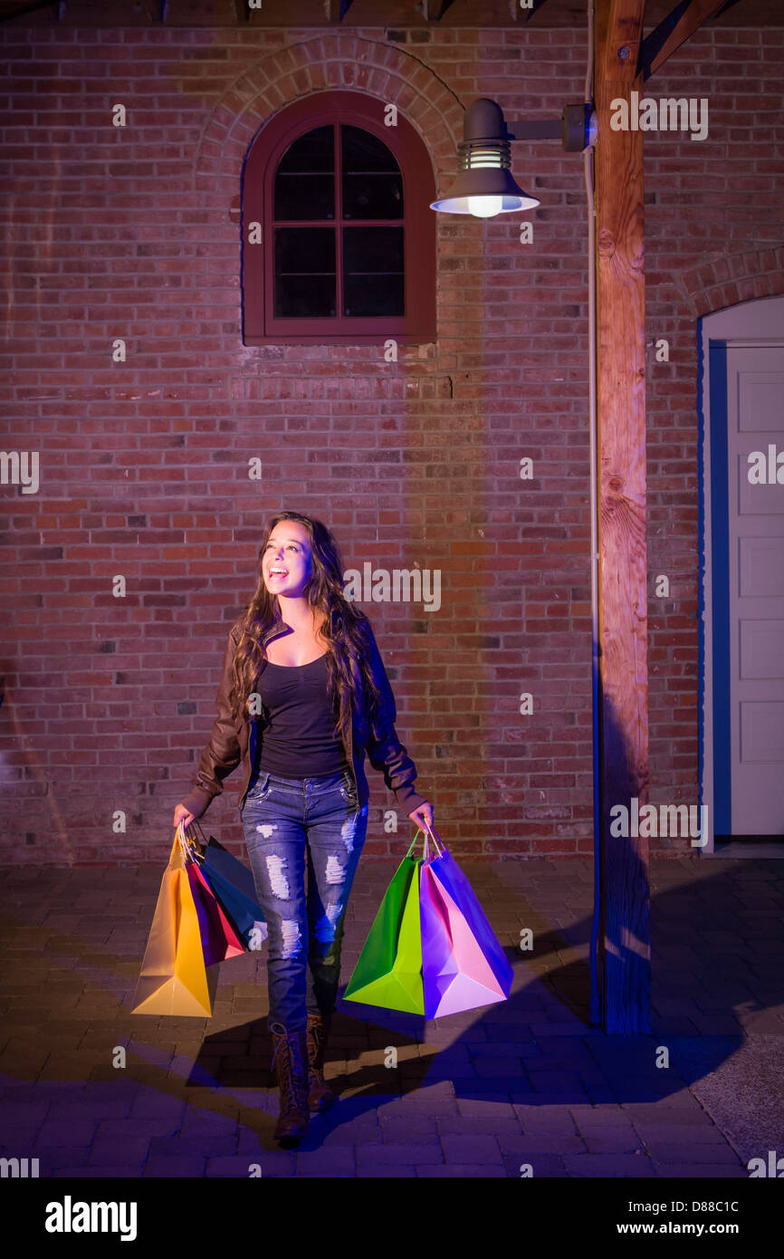 Assez excité Mixed Race Woman Walking in le soir Holding Shopping Bags avec mur de brique Arrière-plan. Banque D'Images