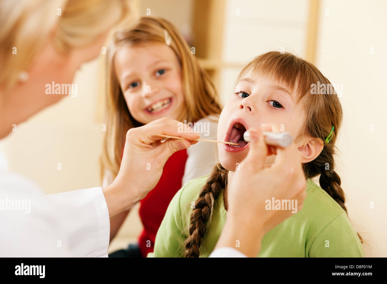Femme médecin pédiatre examinant la gorge d'une jeune fille, dans l'arrière-plan sa soeur est en attente d'être examinés également Banque D'Images
