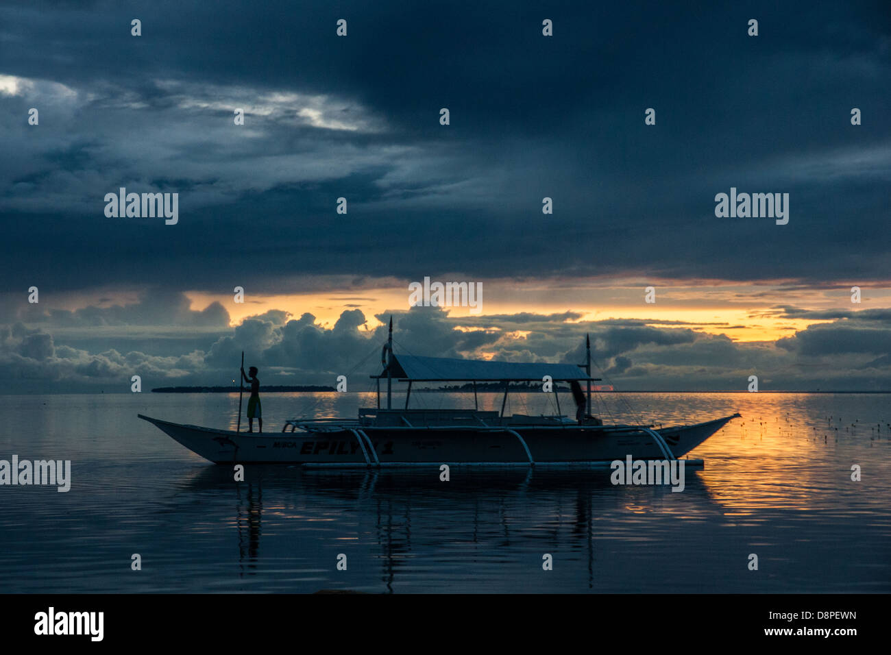 Garçon debout sur la proue de l'outrigger bangka - un bateau de pêche traditionnelle des Philippines - dans le coucher du soleil avec la longue perche dans ses mains Banque D'Images