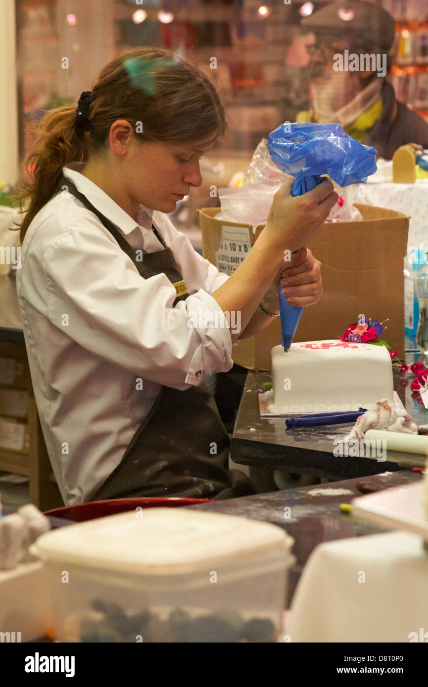 Regarder par la fenêtre de la boutique de gâteau avec une jeune femme avec un sac de tuyauterie à la main décorant le gâteau glacé à Oxford, Oxfordshire Royaume-Uni en mai Banque D'Images