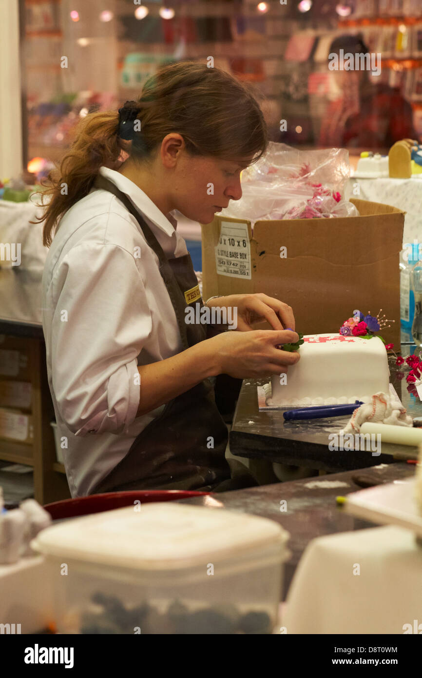 Regarder par la fenêtre de la boutique de gâteau avec une jeune femme décorant le gâteau glacé à Oxford, Oxfordshire Royaume-Uni en mai Banque D'Images