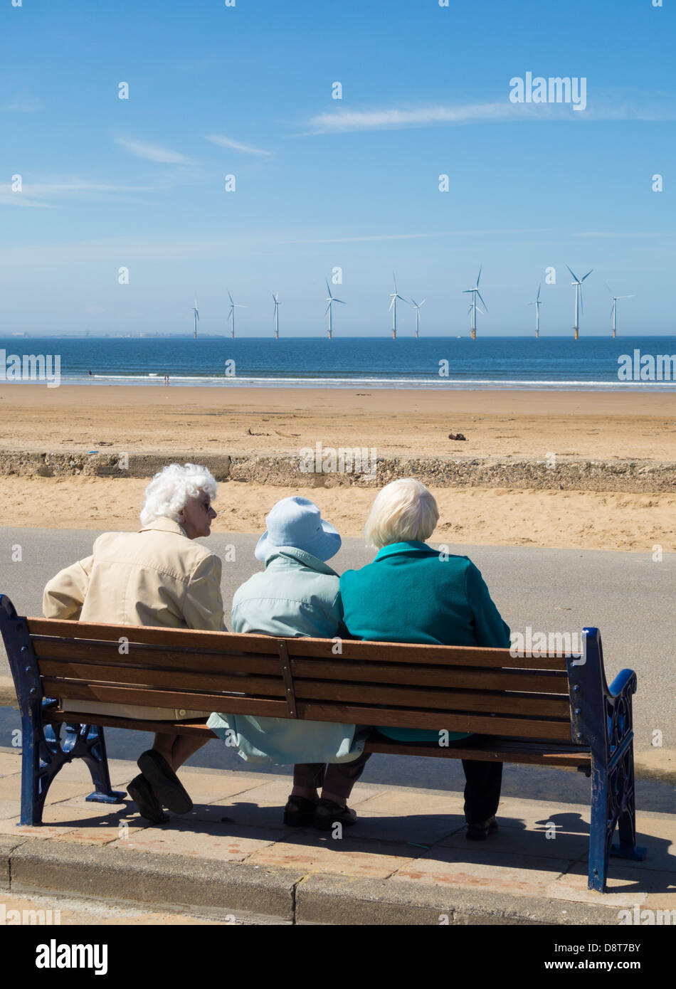 Trois femmes âgées assis sur un banc près de la plage à Redcar, Cleveland, England, UK. Parc éolien offshore de Teesside en arrière-plan Banque D'Images