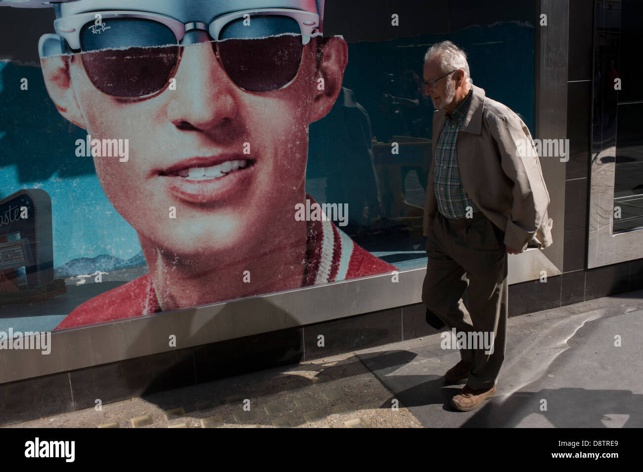 Un monsieur âgé passe par une affiche d'un modèle portant des nuances élégantes à l'extérieur d'une boutique de lunettes de soleil Ray Ban vente fenêtre sur Long Acre in London's Covent Garden. Banque D'Images