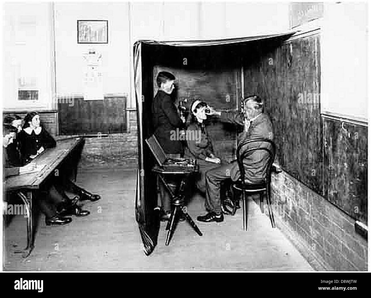 Cette photographie en noir et blanc des archives de State Records NSW montre des enfants subissant des examens de la vue, soulignant les pratiques médicales précoces et l'importance de la santé oculaire. L'image remonte probablement à une époque où de tels tests étaient cruciaux pour assurer le bien-être des enfants dans les milieux éducatifs. Banque D'Images