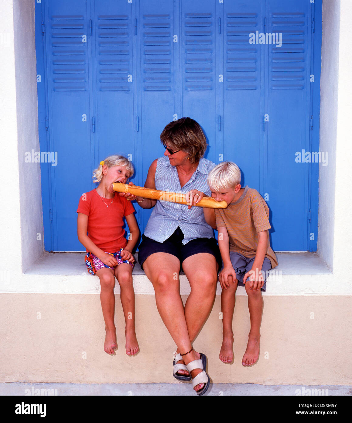 Mère tenant une baguette (Pain français) pour ses enfants, sur un rebord de fenêtre à côté de volets bleus lumineux d'une maison de vacances en Provence, France. Banque D'Images