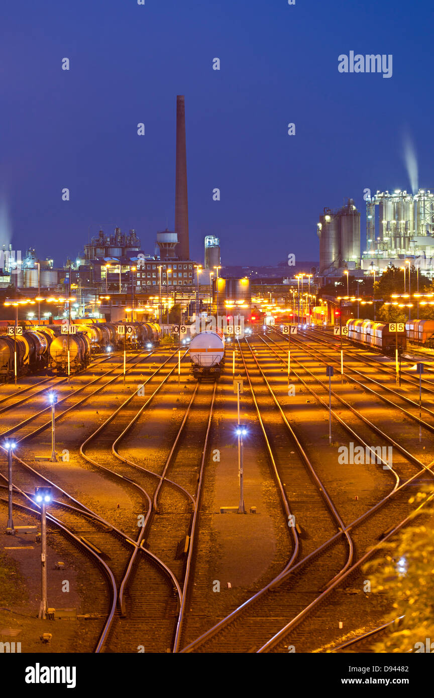 La gare ferroviaire de l'industrie avec l'éclairage orange et bleu ciel de nuit, une usine de produits chimiques dans l'arrière-plan. Banque D'Images