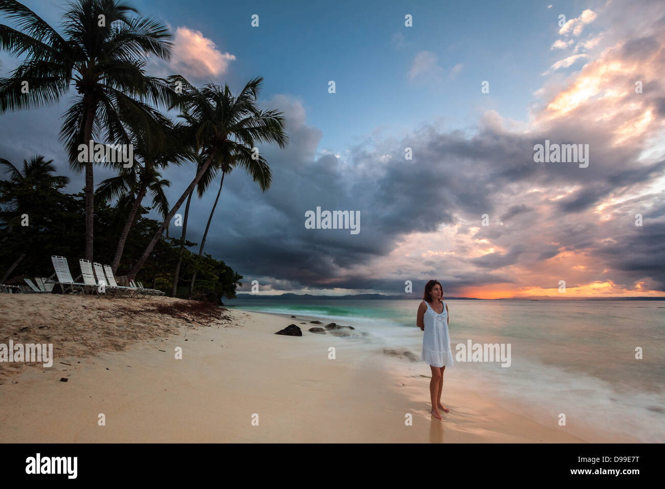 Femme nostalgique dans une robe blanche, pieds nus sur une plage tropicale au coucher du soleil Banque D'Images