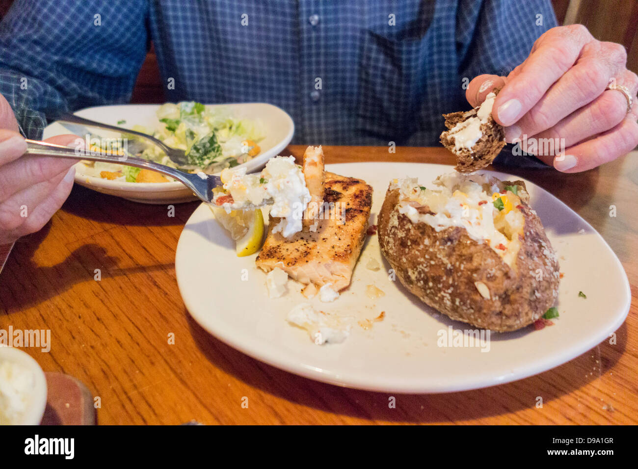 A senior man, la poitrine et les mains seulement, mange un repas de saumon grillé, pomme de terre au four, salade et du pain dans un restaurant. USA Banque D'Images