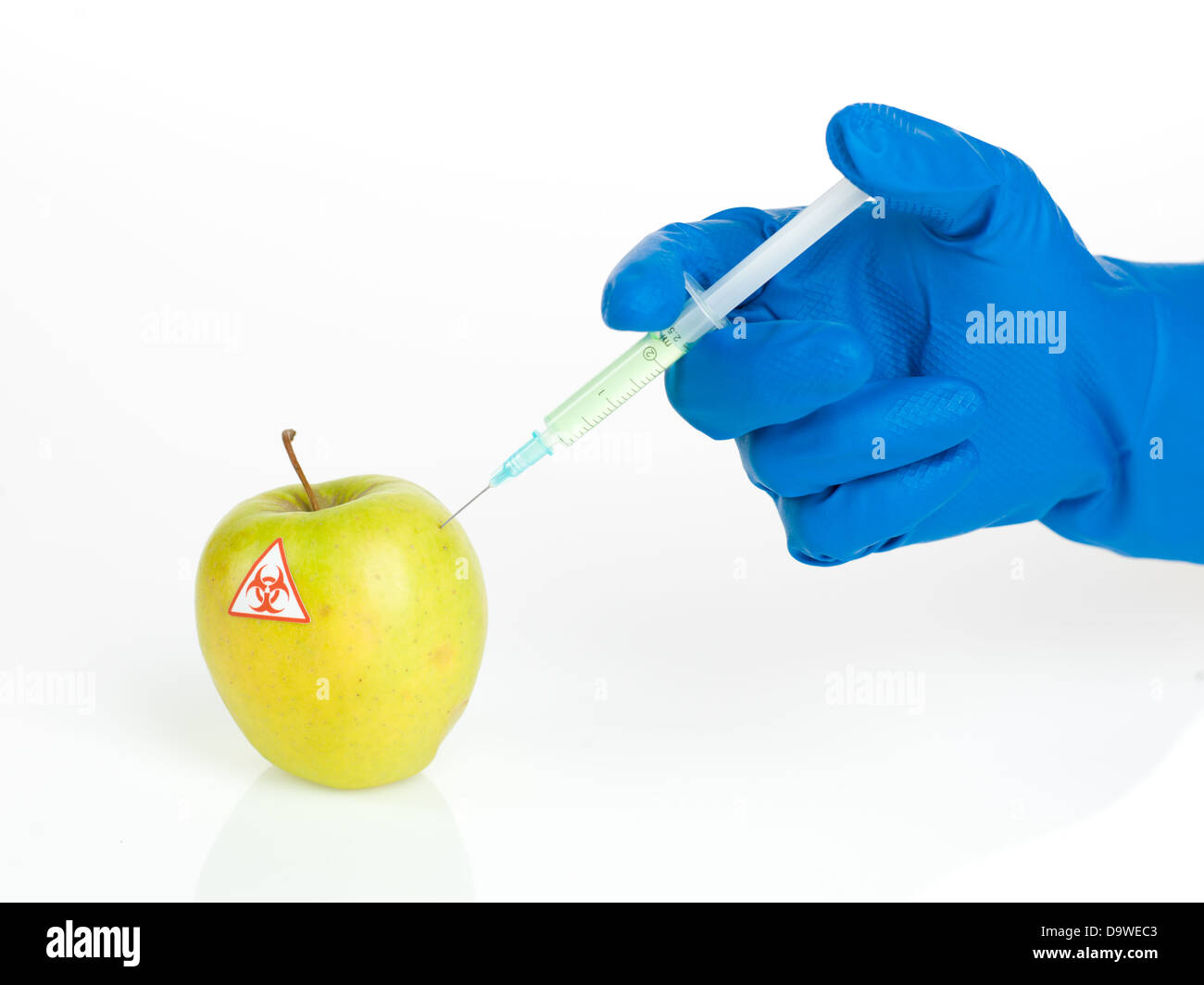 Fond blanc avec une pomme jaune appelée bio être dangereux une injection d'une substance transparente vert clair par une main dans un gant en caoutchouc bleu Banque D'Images