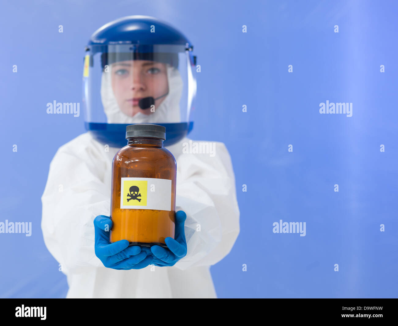 Close-up of female scientist wearing white masque et une combinaison de protection tenant une bouteille étiquetée avec signe mortel, sur bleu backgroun Banque D'Images
