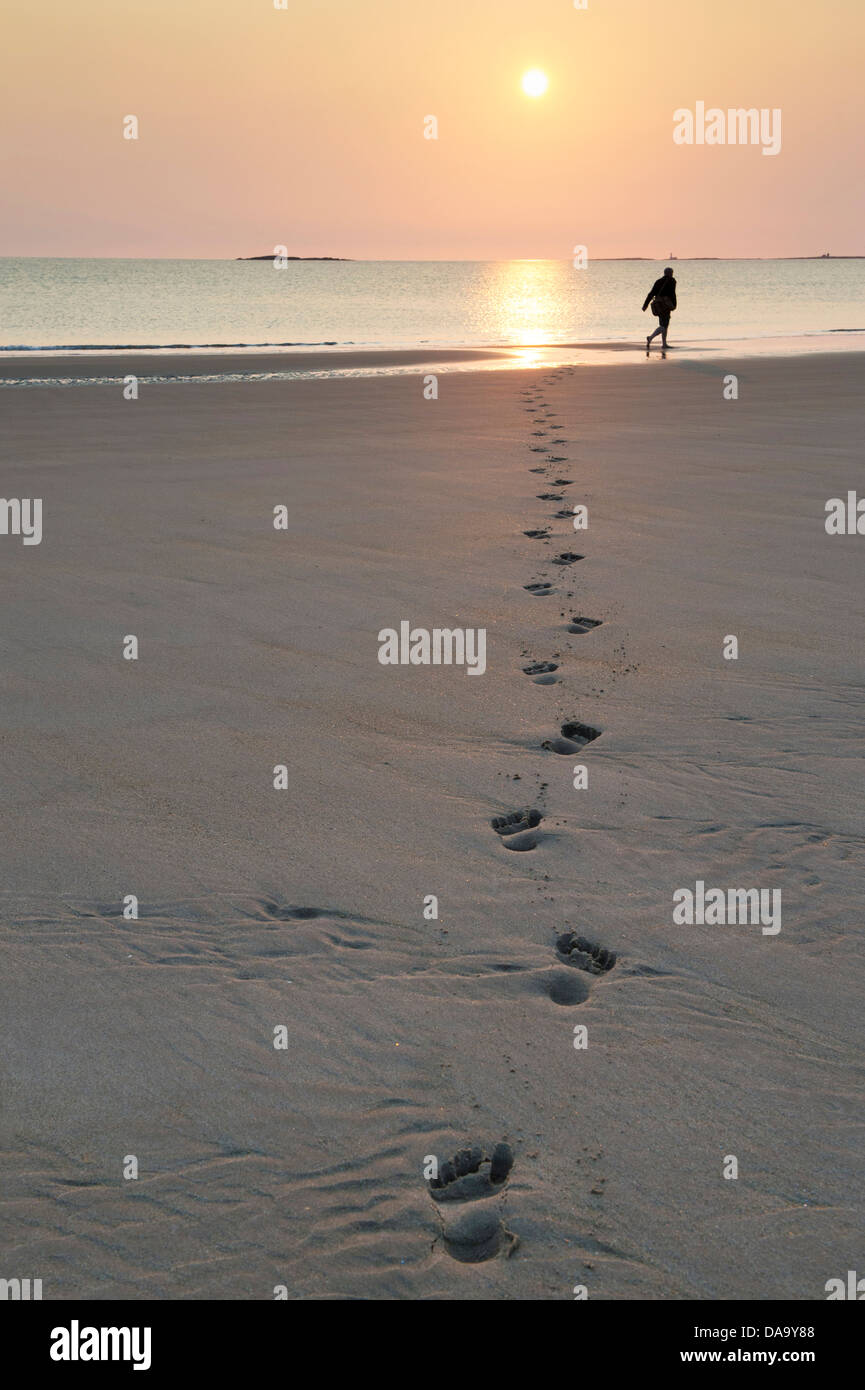 Homme marchant le long de la plage, laissant des traces de pas dans le sable au lever du soleil. , Bamburgh Northumberland, Angleterre Banque D'Images
