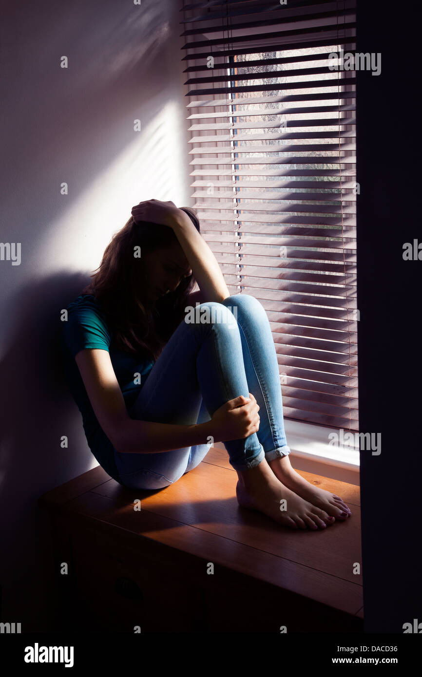 Une adolescente, main sur sa tête, assis par une fenêtre avec la lumière. Banque D'Images