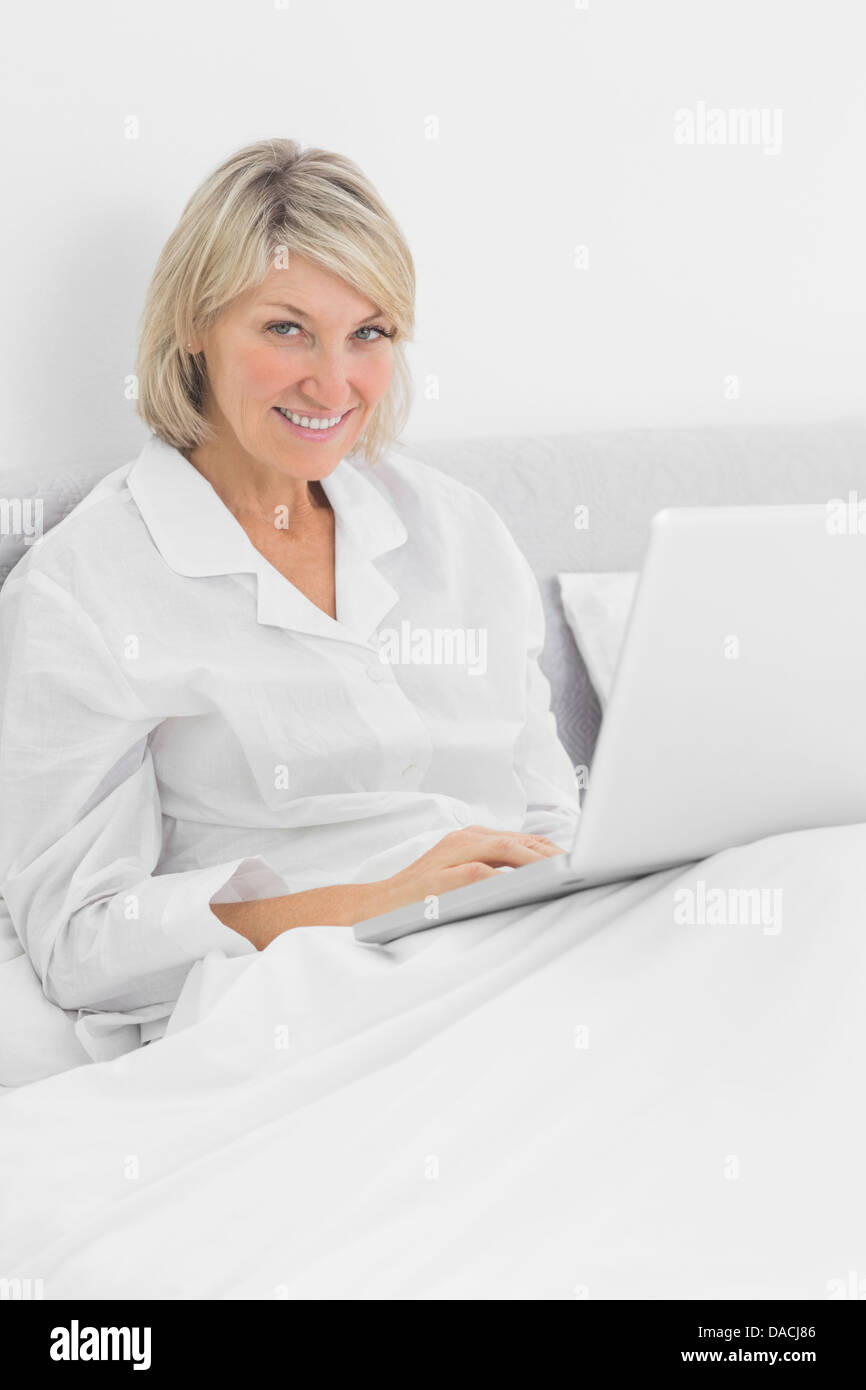 Blonde woman sitting in bed with laptop smiling at camera Banque D'Images