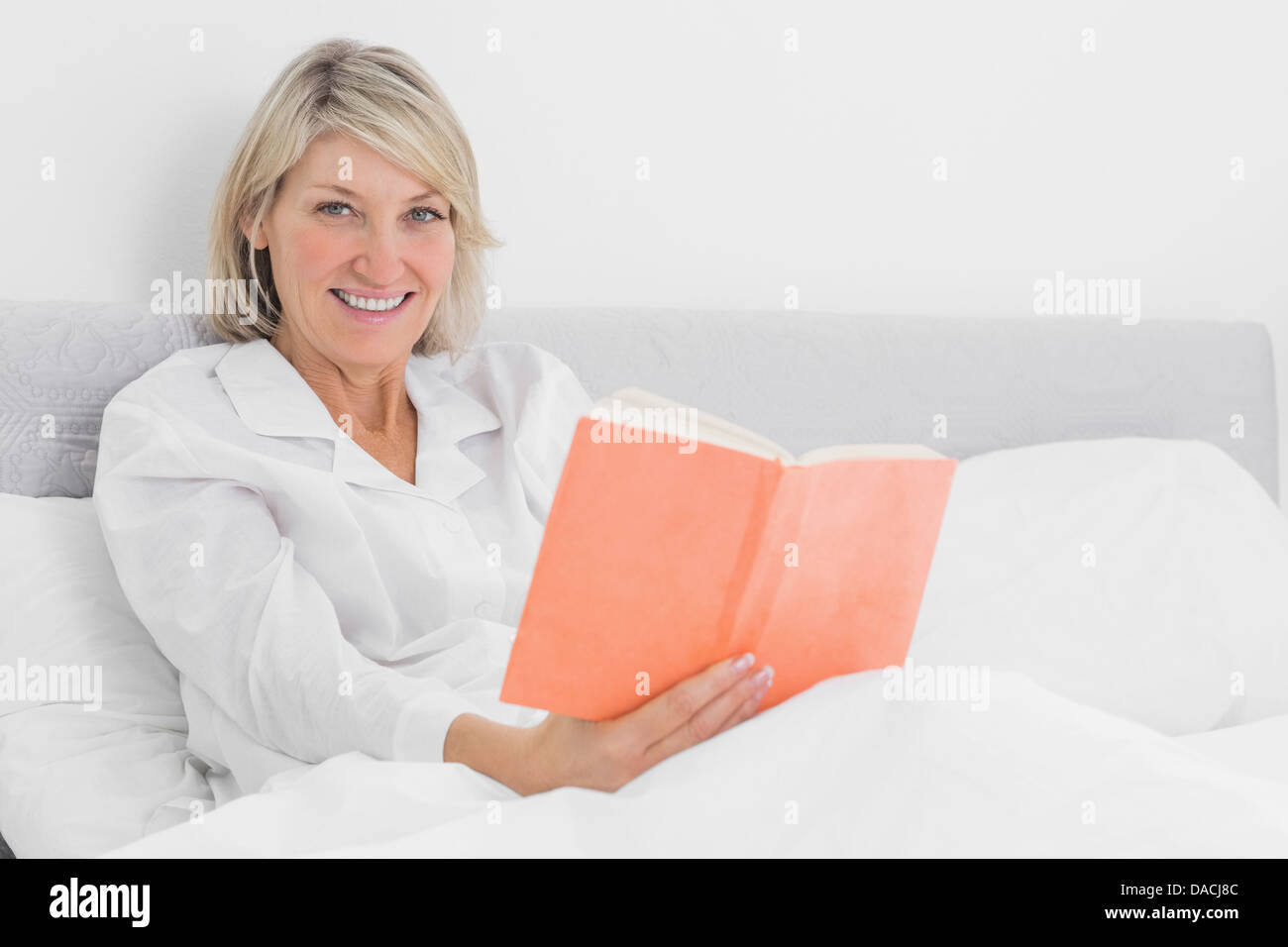 Blonde woman sitting in bed reading smiling at camera Banque D'Images